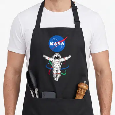 Astronaut nasa apron - Realistic astronaut floating below blue NASA logo with colorful orbital rings — shown on a black cotton canvas apron against a white background
