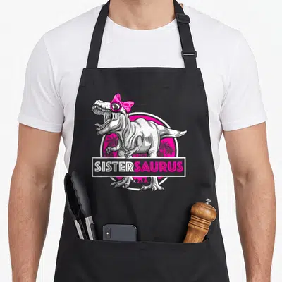 Sistersaurus sister t-rex apron - Cartoon T-Rex with pink bow sunglasses and SISTERSAURUS banner over pink palm circle — shown on a black cotton canvas apron against a white background