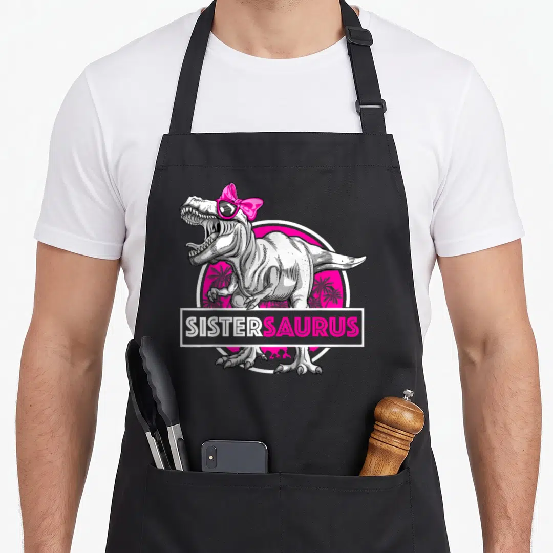 Sistersaurus sister t-rex apron - Cartoon T-Rex with pink bow sunglasses and SISTERSAURUS banner over pink palm circle — shown on a black cotton canvas apron against a white background
