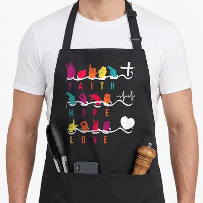 Faith asl sign for apron - Colored ASL hands above ribbons with FAITH HOPE LOVE text and cross heartbeat heart symbols — shown on a black cotton canvas apron against a white background