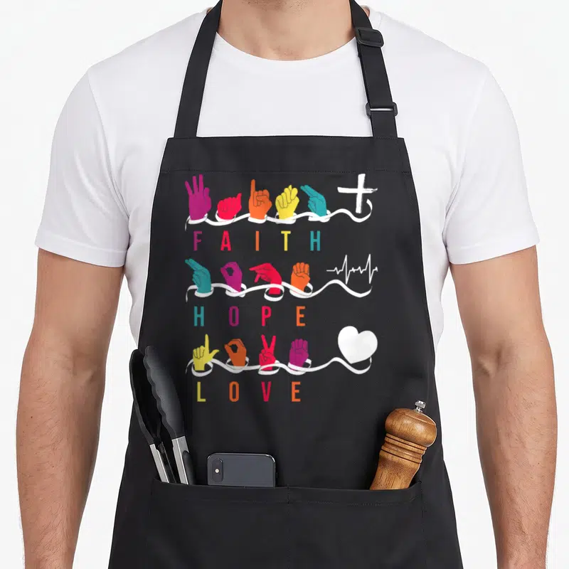 Faith asl sign for apron - Colored ASL hands above ribbons with FAITH HOPE LOVE text and cross heartbeat heart symbols — shown on a black cotton canvas apron against a white background