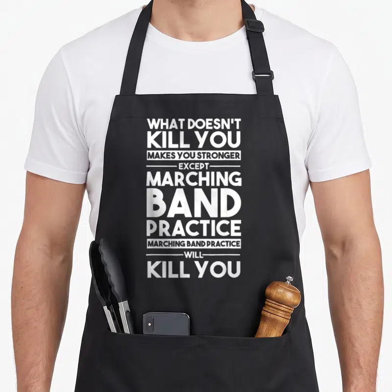 Kill marching band apron - White stacked text reading what doesn't kill you makes you stronger except marching band — shown on a black cotton canvas apron against a white background