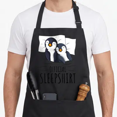 Sleeping penguin aprons - Two cartoon penguins lying on white pillows under a grey blanket with text OFFICIAL — shown on a black cotton canvas apron against a white background