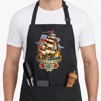 Homeward tattoo apron - Retro sailing ship inside rope oval with HOMEWARD BOUND banner and three flowers below — shown on a black cotton canvas apron against a white background