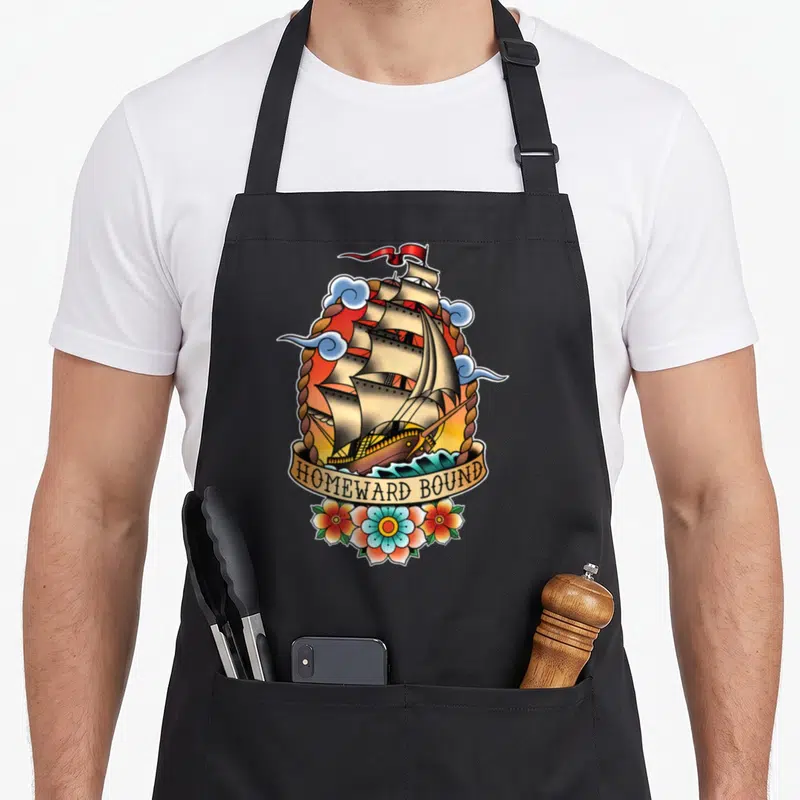 Homeward tattoo apron - Retro sailing ship inside rope oval with HOMEWARD BOUND banner and three flowers below — shown on a black cotton canvas apron against a white background