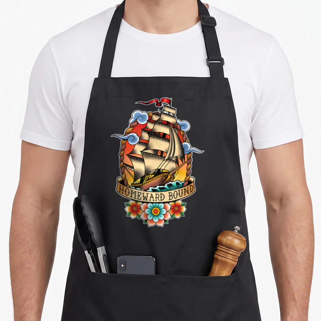 Homeward tattoo apron - Retro sailing ship inside rope oval with HOMEWARD BOUND banner and three flowers below — shown on a black cotton canvas apron against a white background