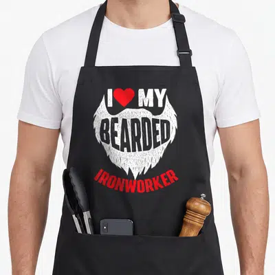 Bearded metal worker apron - White beard graphic with red heart and I MY BEARDED IRONWORKER text in cartoon style — shown on a black cotton canvas apron against a white background