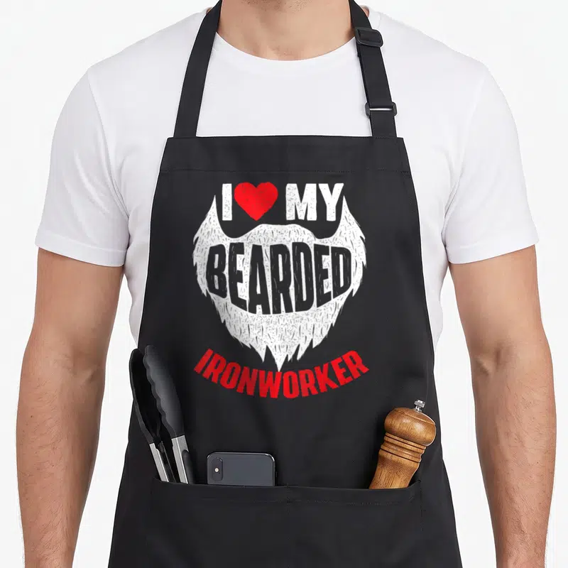 Bearded metal worker apron - White beard graphic with red heart and I MY BEARDED IRONWORKER text in cartoon style — shown on a black cotton canvas apron against a white background
