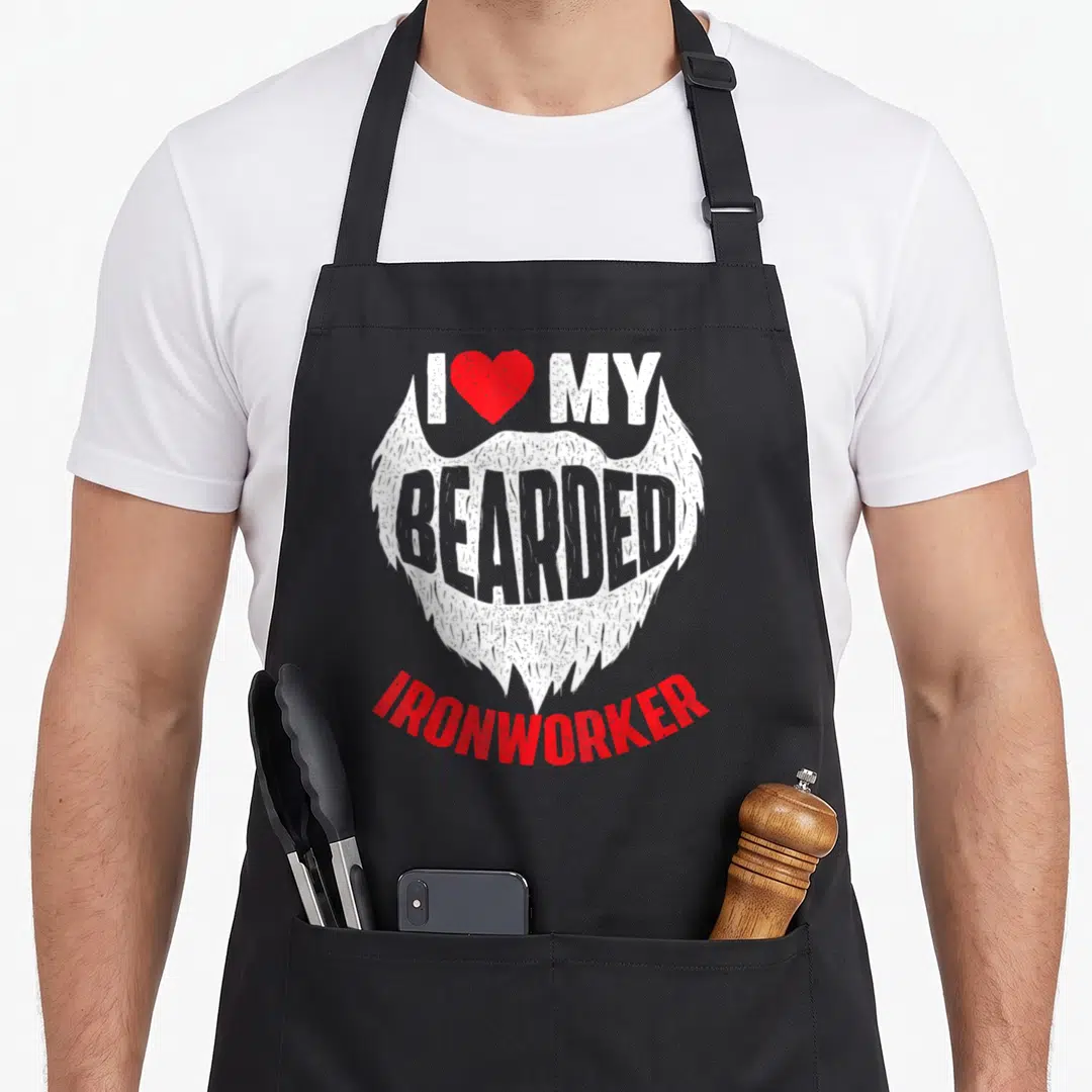 Bearded metal worker apron - White beard graphic with red heart and I MY BEARDED IRONWORKER text in cartoon style — shown on a black cotton canvas apron against a white background