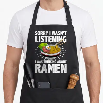 Ramen soup apron - Cartoon ramen bowl with chopsticks and white text saying sorry I wasn't listening I was thinking — shown on a black cotton canvas apron against a white background