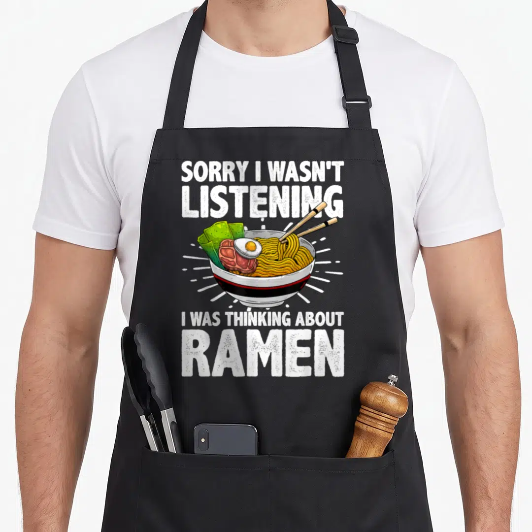 Ramen soup apron - Cartoon ramen bowl with chopsticks and white text saying sorry I wasn't listening I was thinking — shown on a black cotton canvas apron against a white background