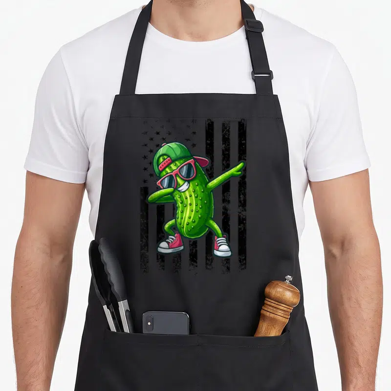 Dabbing american flag apron - Cartoon green pickle dabbing in front of distressed black and grey flag stripes — shown on a black cotton canvas apron against a white background