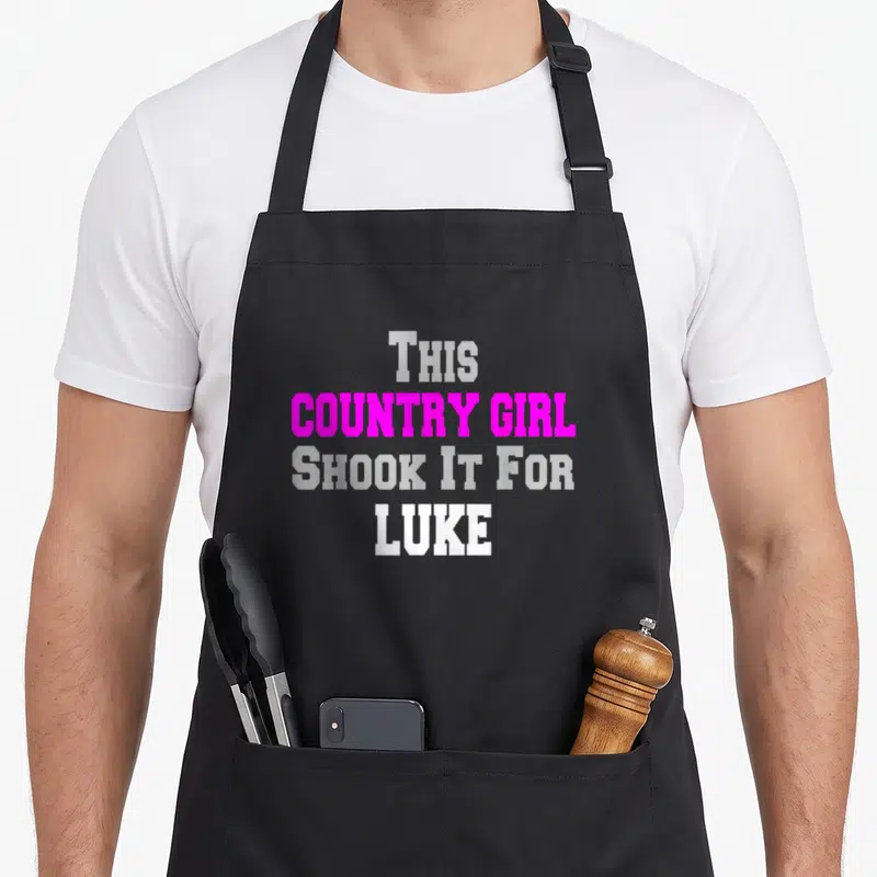 Shook cook's country aprons - Stacked text reading This COUNTRY GIRL SHOOK IT FOR LUKE in magenta grey and white block — shown on a black cotton canvas apron against a white background