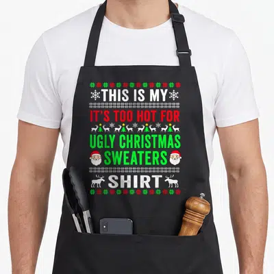 Funny hot ugly christmas apron - Stacked text reading THIS IS MY IT'S TOO HOT FOR UGLY CHRISTMAS SWEATERS SHIRT flanked — shown on a black cotton canvas apron against a white background