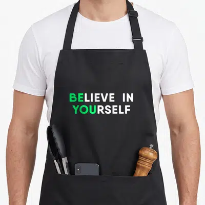 Inspirational motivation apron - BELIEVE IN YOURSELF green and white stacked typography on black background — shown on a black cotton canvas apron against a white background