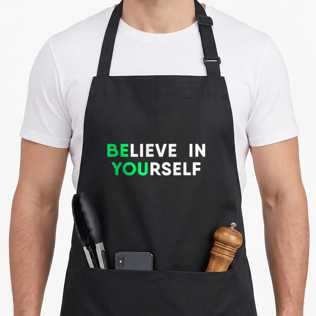 Inspirational motivation apron - BELIEVE IN YOURSELF green and white stacked typography on black background — shown on a black cotton canvas apron against a white background
