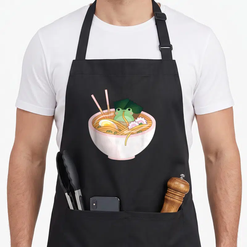 Celestial cartoon apron - Frog sitting in ramen bowl with pink chopsticks and narutomaki in pastel cartoon style — shown on a black cotton canvas apron against a white background