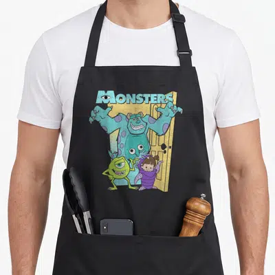 Mike boy monster apron - Sulley in yellow door with Mike and Boo under MONSTERS text — shown on a black cotton canvas apron against a white background