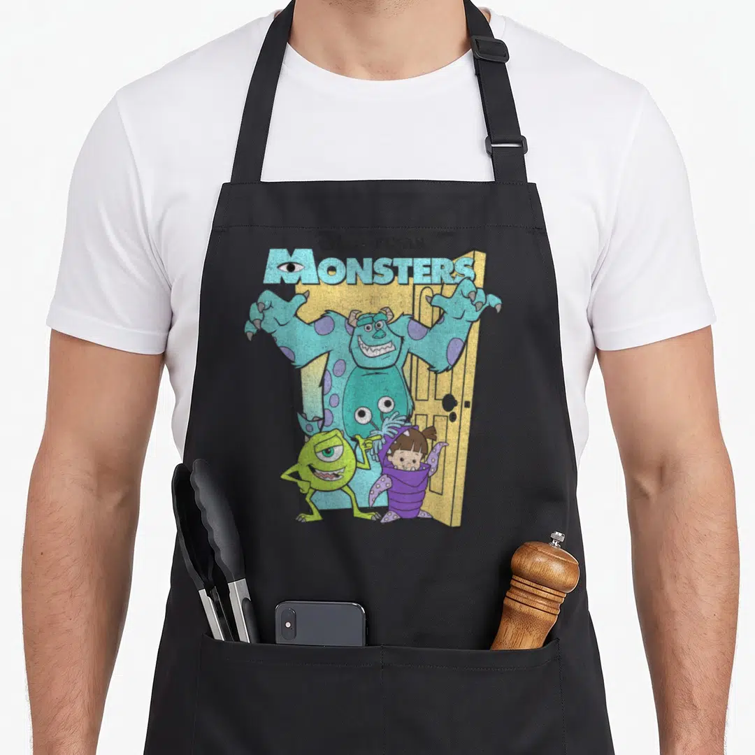 Mike boy monster apron - Sulley in yellow door with Mike and Boo under MONSTERS text — shown on a black cotton canvas apron against a white background