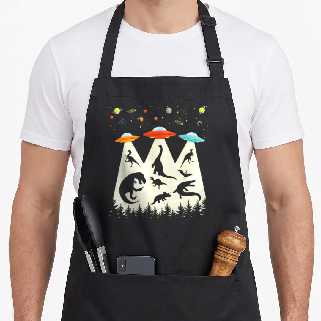 Ufo dinosaur apron - Three UFOs beaming up dinosaur silhouettes above a pine forest line against a grey starry sky — shown on a black cotton canvas apron against a white background