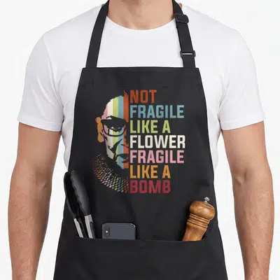 Fragile but apron - Stylized rainbow profile next to stacked text Not Fragile Like A Flower Fragile Like A Bomb — shown on a black cotton canvas apron against a white background