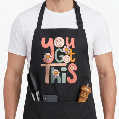 Motivational teacher apron - Stacked YOU GOT THIS text with retro smiley faces and flowers in pink orange blue grey — shown on a black cotton canvas apron against a white background