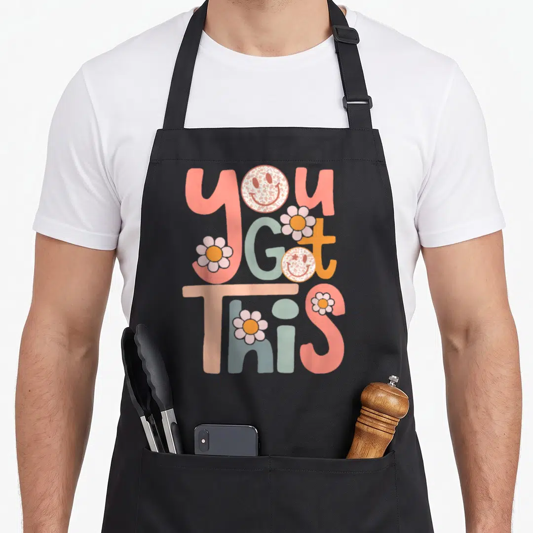 Motivational teacher apron - Stacked YOU GOT THIS text with retro smiley faces and flowers in pink orange blue grey — shown on a black cotton canvas apron against a white background