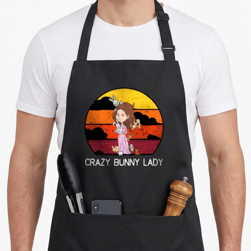 Cute bunny rabbit apron - Cartoon woman holding rabbits inside yellow orange red circle with black clouds and white — shown on a black cotton canvas apron against a white background