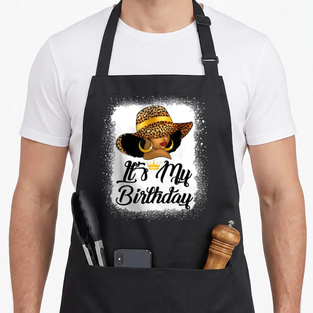 African queen apron - Cartoon woman in leopard hat with gold crown and It's My Birthday text — shown on a black cotton canvas apron against a white background