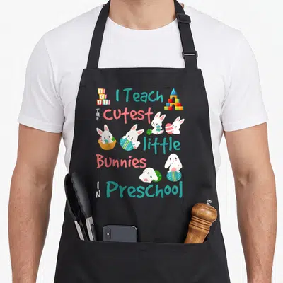 Easter preschool teacher apron - Cartoon white bunnies holding Easter eggs with ABC blocks and colorful text — shown on a black cotton canvas apron against a white background