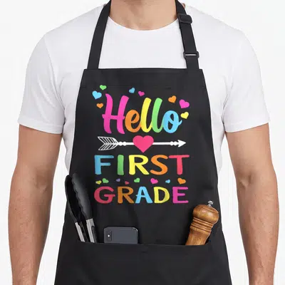 1st teacher apron - Colorful Hello First Grade text with pierced heart and scattered multicolored hearts — shown on a black cotton canvas apron against a white background
