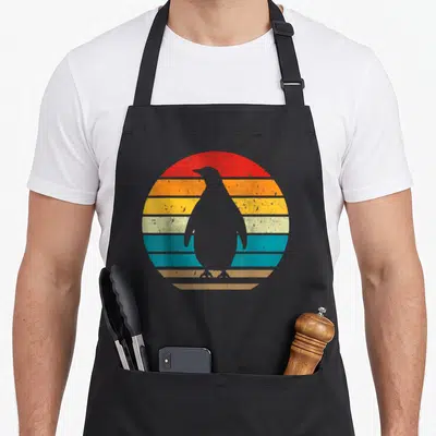 Sunset penguin aprons - Minimalist penguin silhouette against a striped retro sunset in red, orange, yellow, teal — shown on a black cotton canvas apron against a white background
