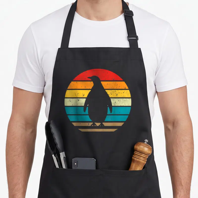 Sunset penguin aprons - Minimalist penguin silhouette against a striped retro sunset in red, orange, yellow, teal — shown on a black cotton canvas apron against a white background
