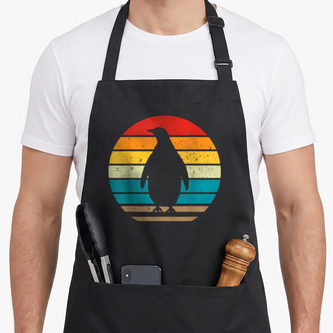 Sunset penguin aprons - Minimalist penguin silhouette against a striped retro sunset in red, orange, yellow, teal — shown on a black cotton canvas apron against a white background