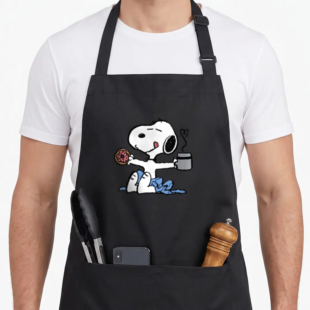 Funny coffee donut apron - Snoopy in blue blanket holding pink donut and grey mug with heart steam — shown on a black cotton canvas apron against a white background