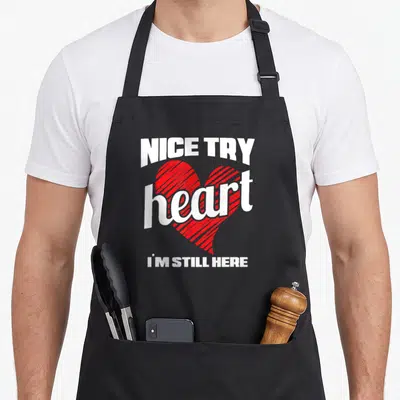 Attack distillery apron - NICE TRY I'M STILL HERE text surrounding a red striped heart graphic — shown on a black cotton canvas apron against a white background