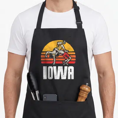 Iowa wrestler wrestling apron - Retro graphic of two wrestlers grappling before striped sun with white IOWA text below — shown on a black cotton canvas apron against a white background