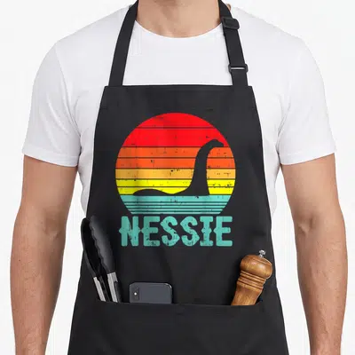 Nessie animal apron - Retro Nessie silhouette in striped red orange yellow teal sun with text — shown on a black cotton canvas apron against a white background