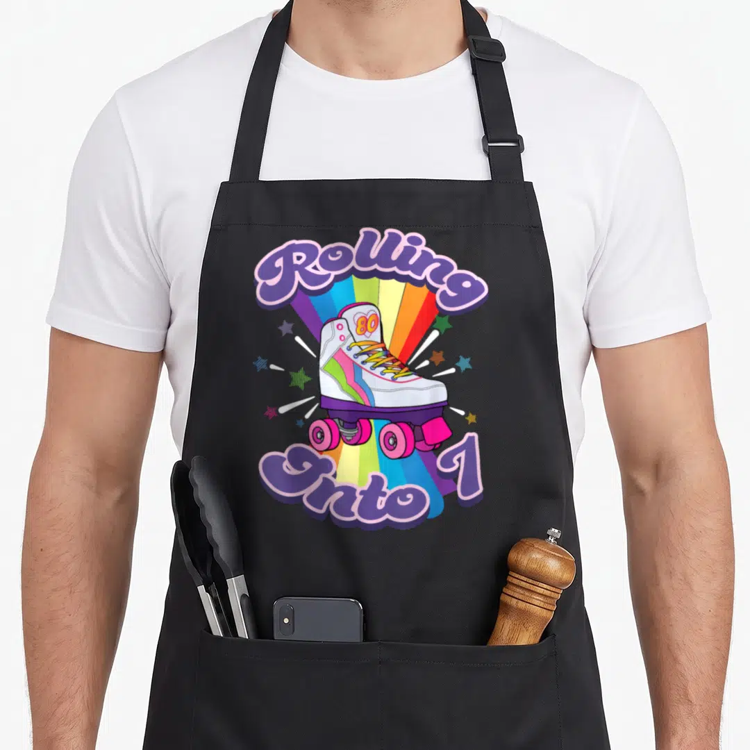 Skating 7th birthday apron - Rolling Into 1 text surrounding a white roller skate on a rainbow burst background — shown on a black cotton canvas apron against a white background