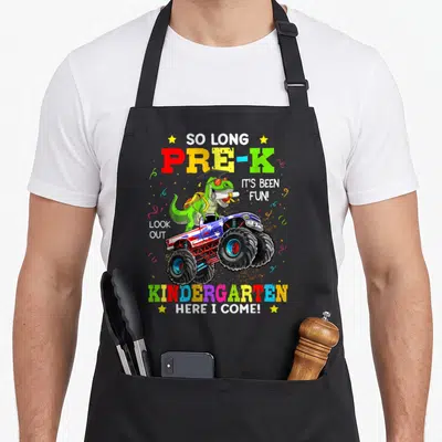 Dinosaur come and get it apron - Green dinosaur in monster truck with Pre-K and Kindergarten graduation text — shown on a black cotton canvas apron against a white background