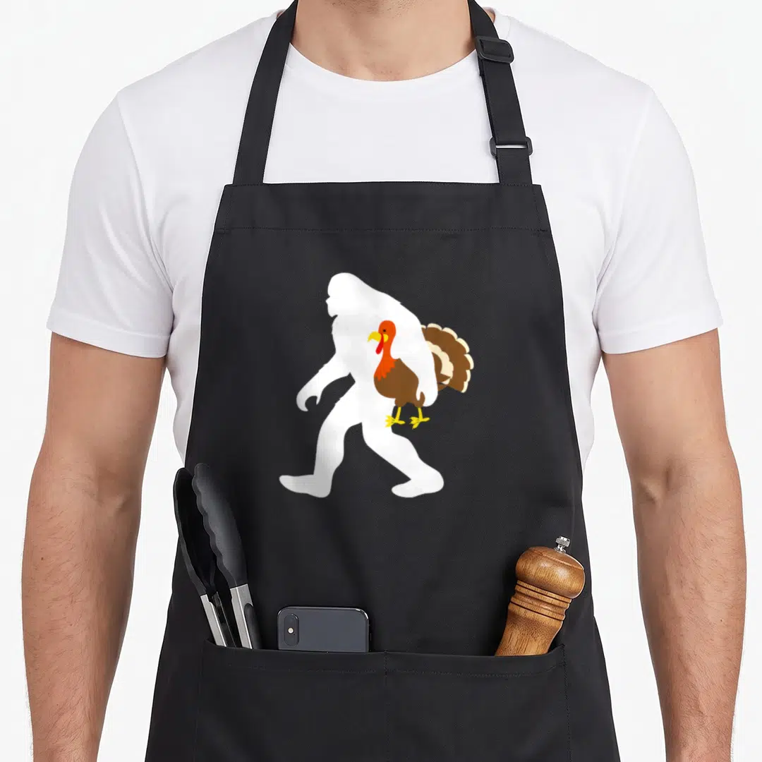 Bigfoot turkey apron - White silhouette walking left carrying brown turkey with red wattle — shown on a black cotton canvas apron against a white background