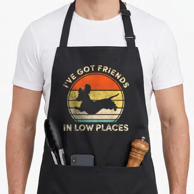 Friend wiener apron - Grey dachshund silhouette over orange yellow striped sun with text I've Got Friends In Low Places — shown on a black cotton canvas apron against a white background