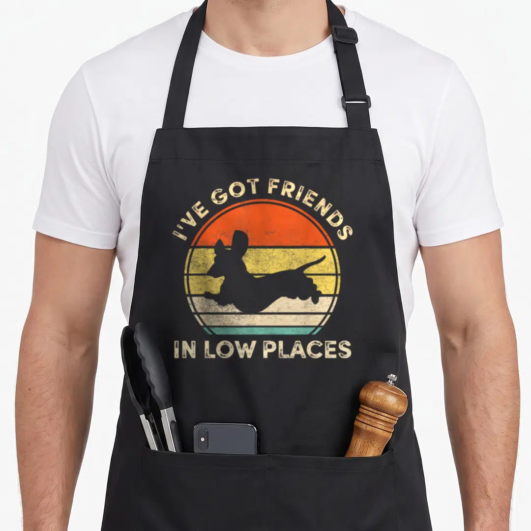 Friend wiener apron - Grey dachshund silhouette over orange yellow striped sun with text I've Got Friends In Low Places — shown on a black cotton canvas apron against a white background
