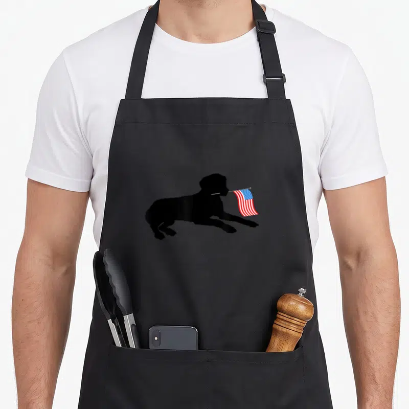 Lab 4th of july apron - Black silhouette of a leaping dog holding a small waving USA flag in its mouth — shown on a black cotton canvas apron against a white background
