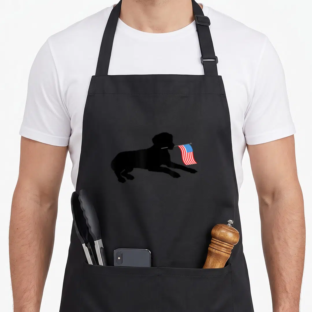 Lab 4th of july apron - Black silhouette of a leaping dog holding a small waving USA flag in its mouth — shown on a black cotton canvas apron against a white background