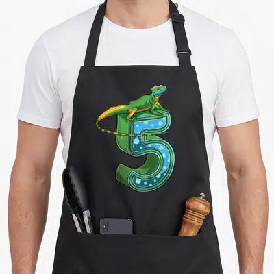 Lizard birthday apron - Cartoon iguana perched on 3D number 5 with green blue and yellow details — shown on a black cotton canvas apron against a white background