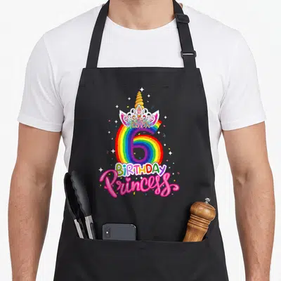6th princess apron - Rainbow number 6 with unicorn horn, tiara, BIRTHDAY text, and pink Princess script surrounded — shown on a black cotton canvas apron against a white background