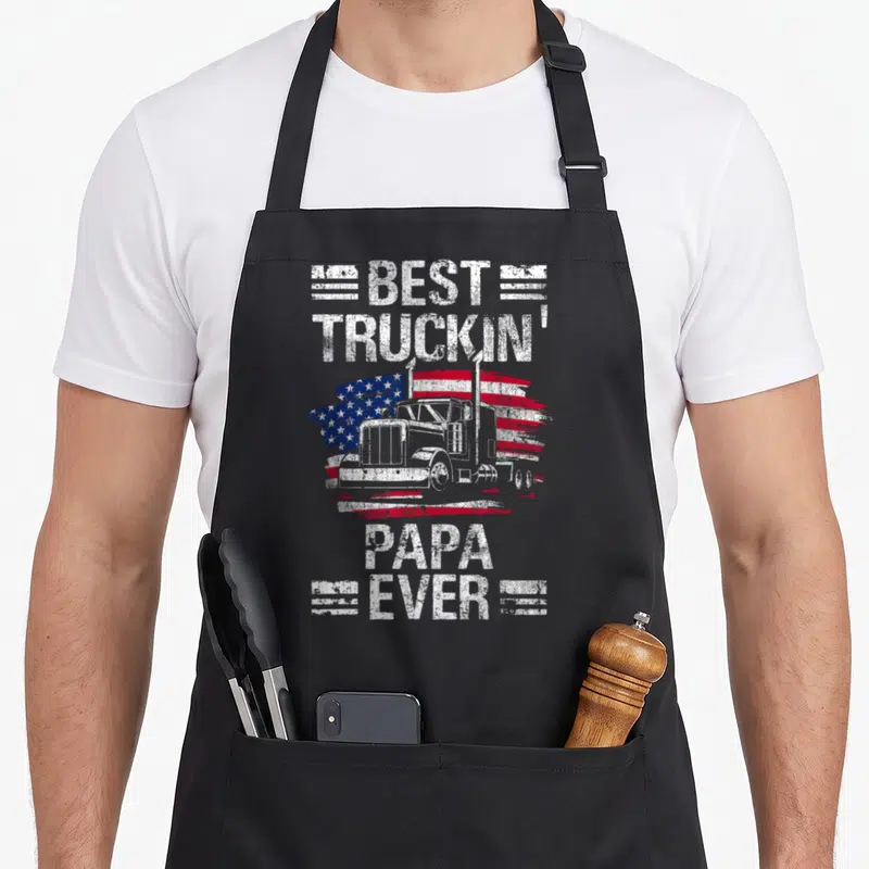 Flag semi truck apron - Best Truckin Papa Ever text with semi-truck and distressed USA flag graphic — shown on a black cotton canvas apron against a white background