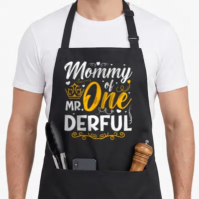 Derful family aprons - Stacked white and yellow text reading Mommy of Mr One Derful with crown and hearts — shown on a black cotton canvas apron against a white background