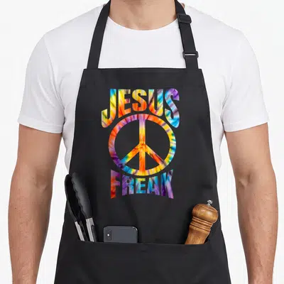 Jesus tie-dye apron - Arched JESUS FREAK text framing a peace symbol in red yellow blue purple tie-dye pattern — shown on a black cotton canvas apron against a white background